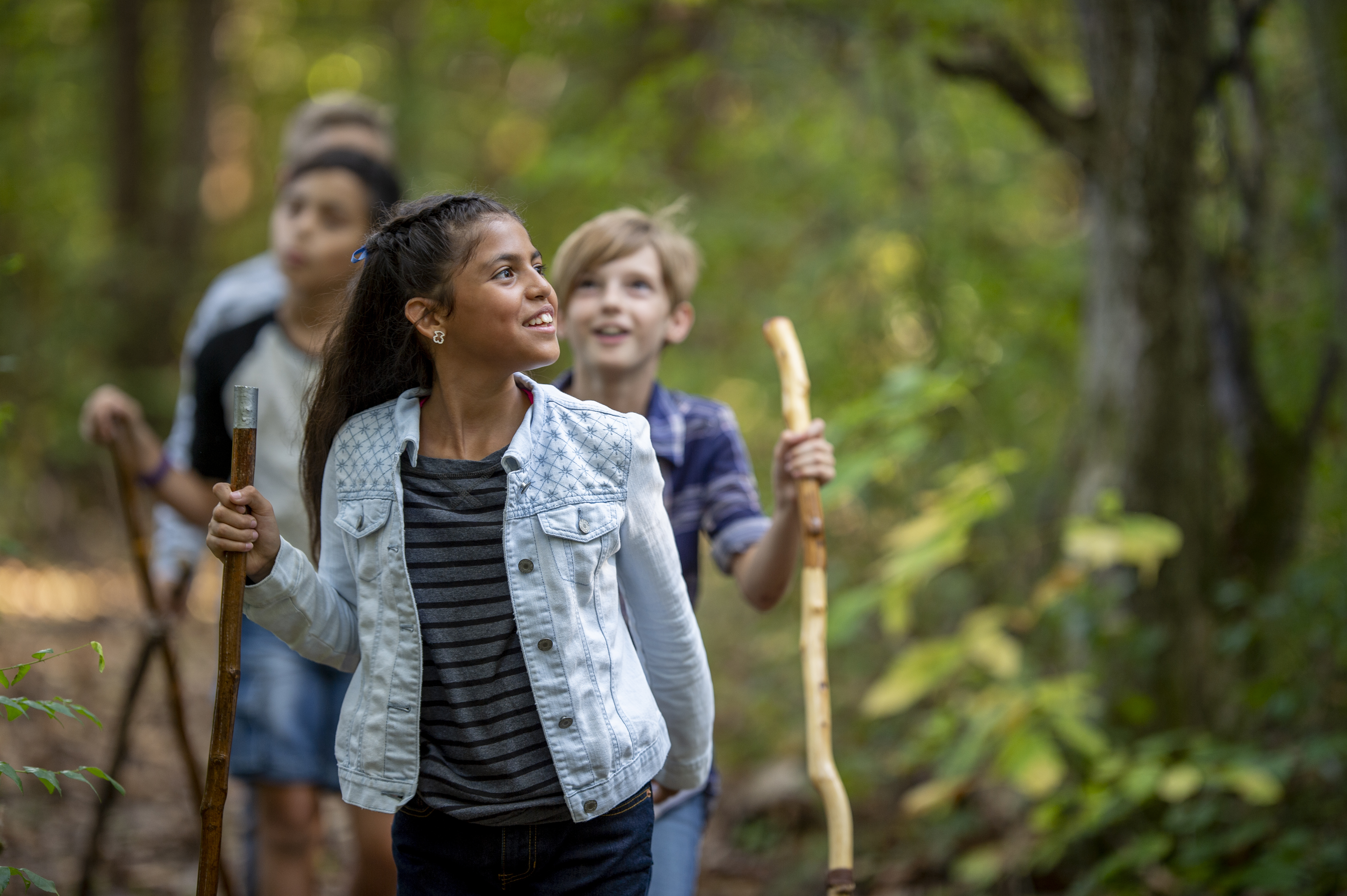 Four children walk in a single file line through a forest scene. They are each holding a walking stick. The first child in line is fully in focus, wearing a light wash denim jacket, dark grey striped shirt, and earrings. They are smiling at the forest in awe and have deep brown skin, brown eyes, and dark hair tied off their face with a blue ribbon. The children behind them are all slightly out of focus but have the same excited smiles.