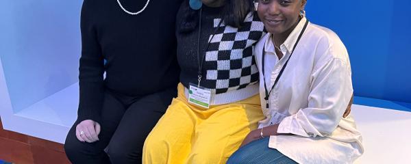Three individuals sit smiling at the camera in front of a blue background. The first from the left has red hair, wears glasses and is in a black shirt and pants. A dark-skinned Black person with long hair is sitting in the middle wearing a black and white checker-patterned top and yellow skirt. The person on the right of the image is sitting with their legs crossed and has short hair and is wearing a white top and blue pants. 