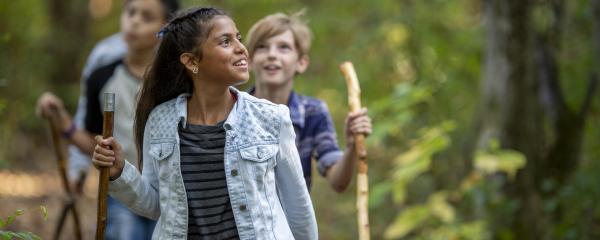 Four children walk in a single file line through a forest scene. They are each holding a walking stick. The first child in line is fully in focus, wearing a light wash denim jacket, dark grey striped shirt, and earrings. They are smiling at the forest in awe and have deep brown skin, brown eyes, and dark hair tied off their face with a blue ribbon. The children behind them are all slightly out of focus but have the same excited smiles.