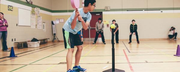 Students in a gymnasium participate in a variety of activities. The focus of the image is a student preparing to swing a racquet at a tennis ball mounted on a tee. Three students wait to catch the ball some distance away. 