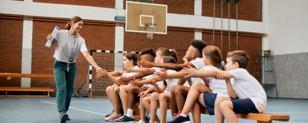 A feminine-presenting gym teacher high fives a line of students extending their hands while sitting on a bench in a gymnasium. The teacher has long brown hair that is tied back in a ponytail, and is wearing athletic clothing and holding a clipboard. The students are diverse in terms of gender expression and skin tone. Everyone is smiling enthusiastically.