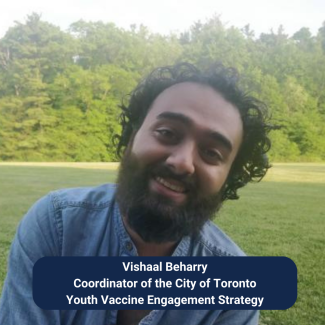 Blog contributor Vishaal Beharry, a South Asian man, smiles as he sits on the ground in a field of green grass. He wears a beard, and has dark short curly hair. He is wearing a denim shirt.