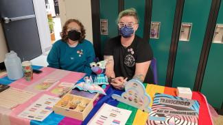 ""Image of two individuals wearing Kn95 masks seated at a table in front of a row of green lockers. The person on the left has short red curly hair, glasses, and wears a teal crewneck decorated with pins and stickers featuring the rainbow flag. The person on the right has short hair with blue tips that is styled in a combover. They wear a black t-shirt and have tattoos on both arms. The table they are both seated at features resources from the CCGSD about 2SLGBTQIA+ identities. 