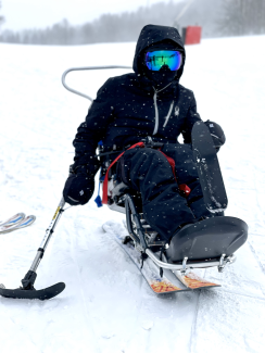 Jake Wttewaal on a snowy ski slope using an adaptive bi-ski. He is wearing a black ski jacket and ski pants, with blue mirrored ski goggles, a black balaclava, and black ski gloves. His ski poles rest on the ground beside him. 