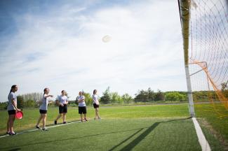 Cinq élèves en tenue de sport sont debout sur un terrain devant un filet de soccer. L'un d'entre eux vient de lancer un frisbee blanc, que les élèves regardent voler vers le filet. 