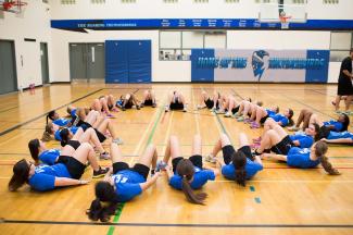 Un grand groupe d'élèves allongés sur le dos, formant un grand cercle avec leurs jambes pointées vers l'intérieur sur le sol d'un gymnase. Les élèves portent des vêtements de sport assortis, avec des t-shirts bleus, et la plupart d'entre eux ont les cheveux longs attachés en queue de cheval. 