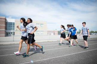 Des élèves courent et marchent à différentes allures sur une piste d'athlétisme extérieure. Les élèves portent des uniformes de sport assortis et semblent courir devant une école. 