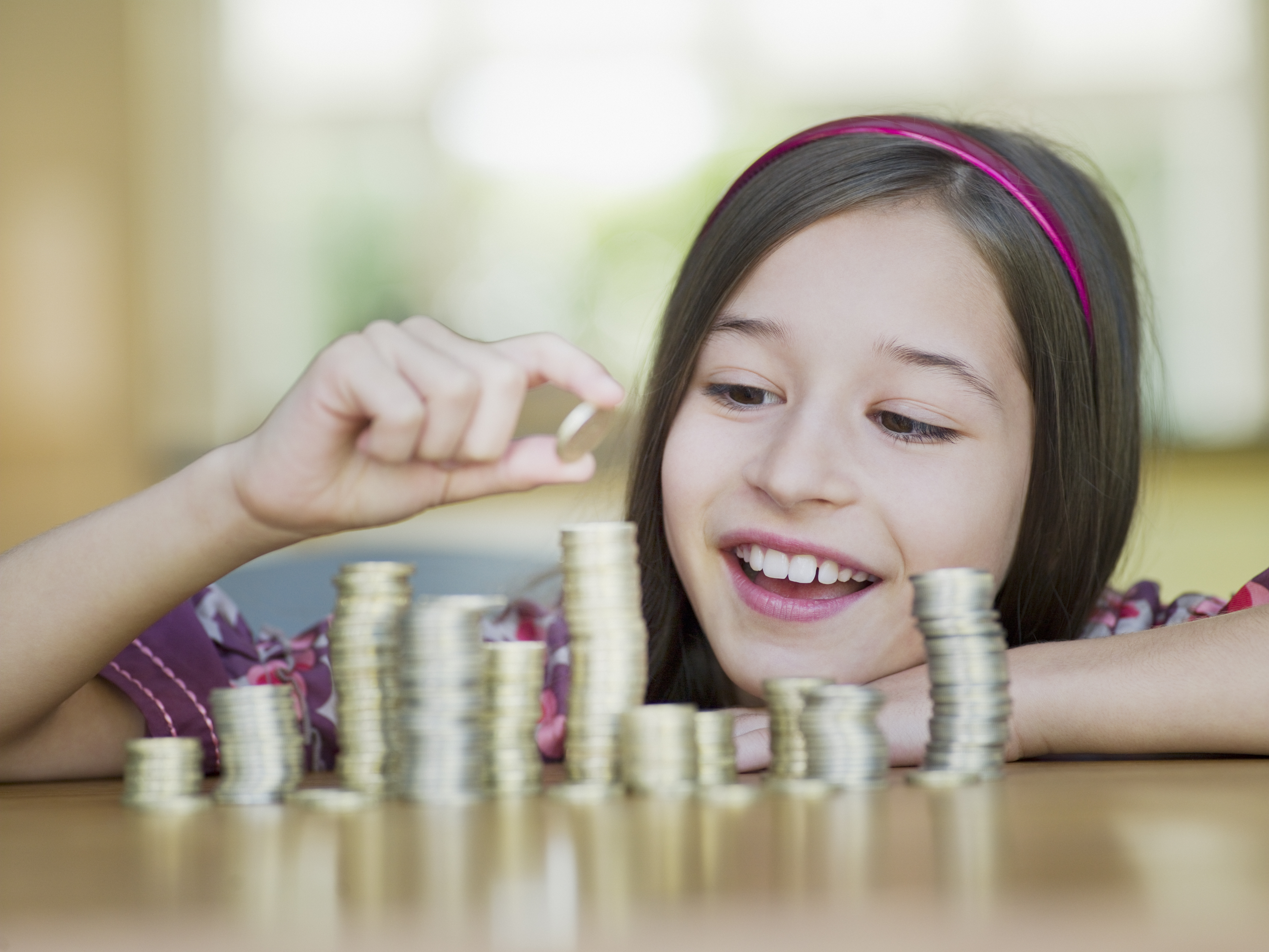 A young child with long hair rests their forearms on a table with piles of different coins. The piles are different sizes, and the child is smiling while placing a coin at the top of the highest pile.