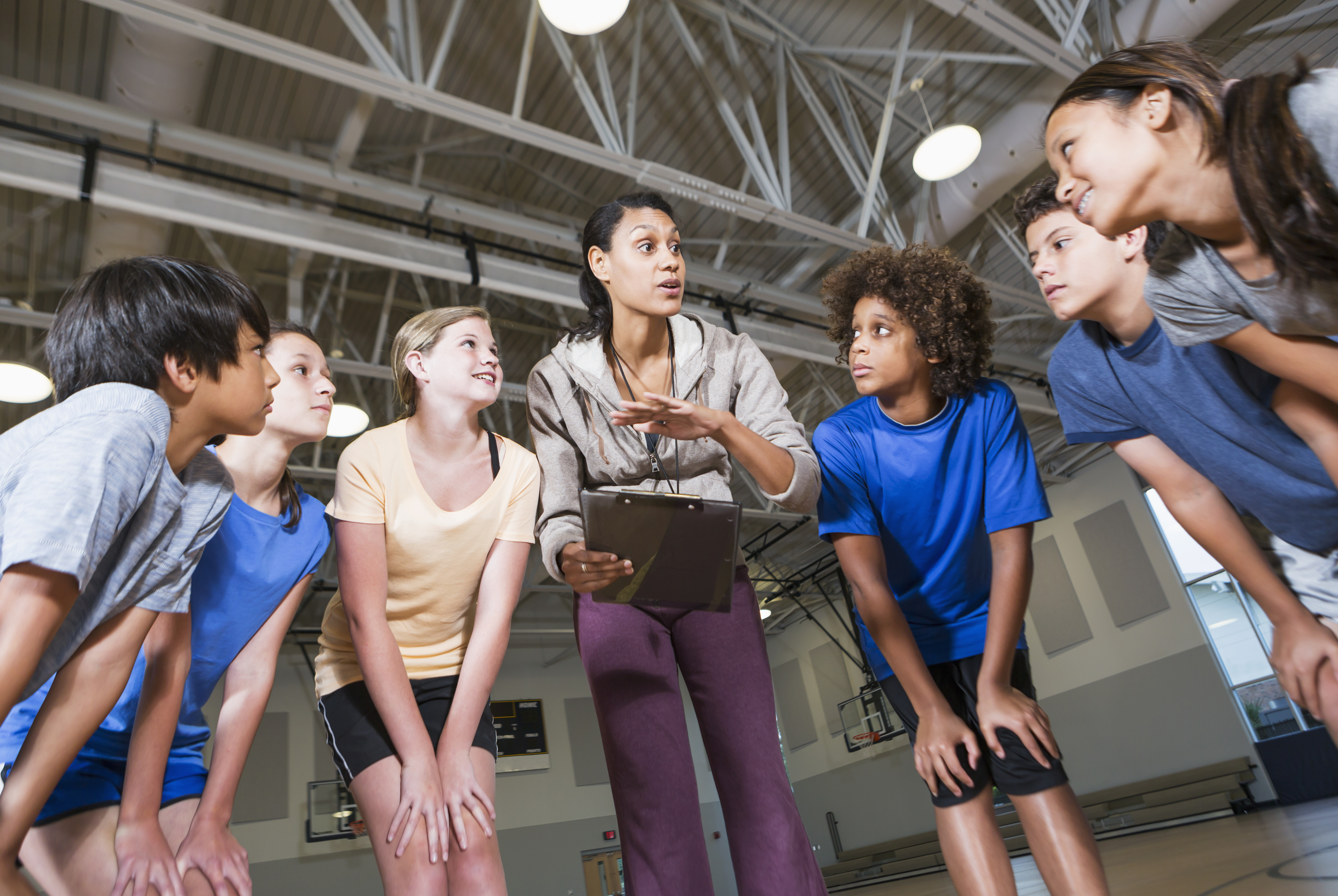 A teacher and a group of students stand in a huddle in the middle of a gymnasium. The teacher is holding a clipboard and speaking to the students, who are listening intently. Everyone in the photo is wearing athletic clothing.