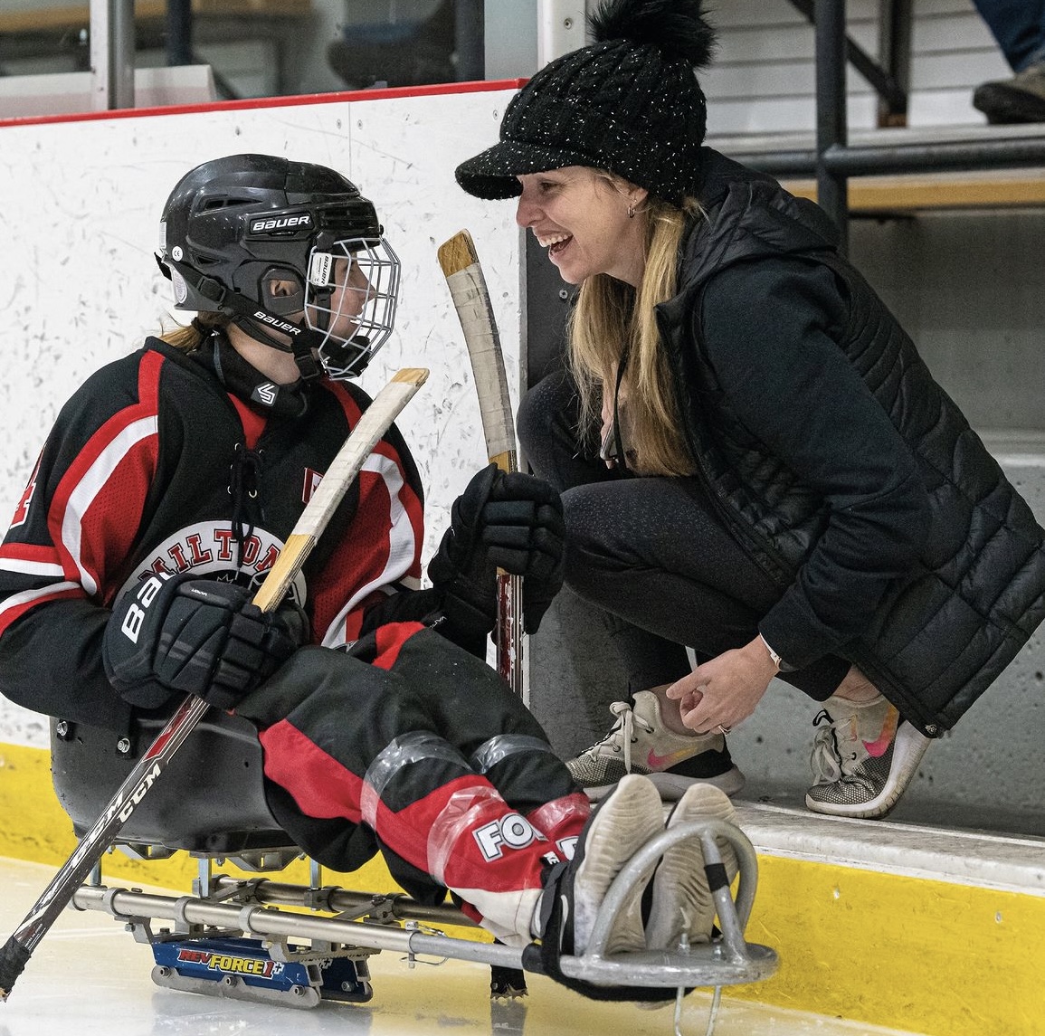 Jennifer Gallupe Roos, a white woman with long blonde hair, kneels at the open door of a bench at a hockey arena beside a young athlete in a sledge-hockey sledge. Jenn is wearing a black hat and black jacket, and the student wears a helmet and red and black gear.