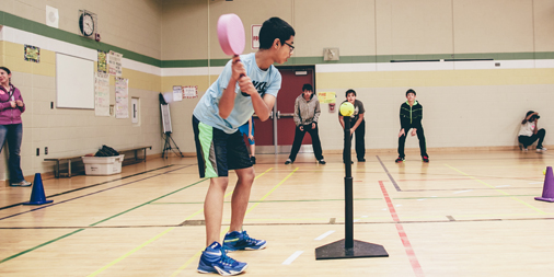 Students in a gymnasium participate in a variety of activities. The focus of the image is a student preparing to swing a racquet at a tennis ball mounted on a tee. Three students wait to catch the ball some distance away. 