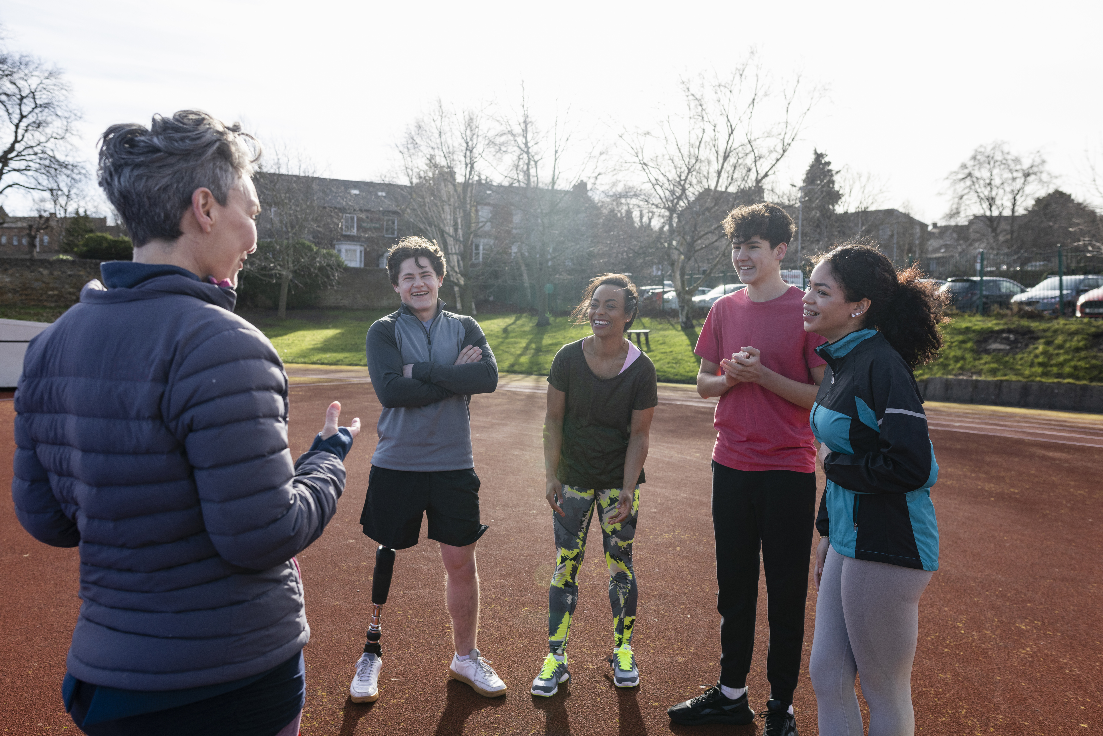 Four students stand facing their teacher on an outdoor track in late Fall. The teacher, who has short grey hair and wears a grey puffer jacket, has their back turned and is gesturing with their hands. All of the students wear athletic clothes, and they display a range of skin tones, gender expressions, and hair textures. One of the students is an amputee and has a prosthetic leg.