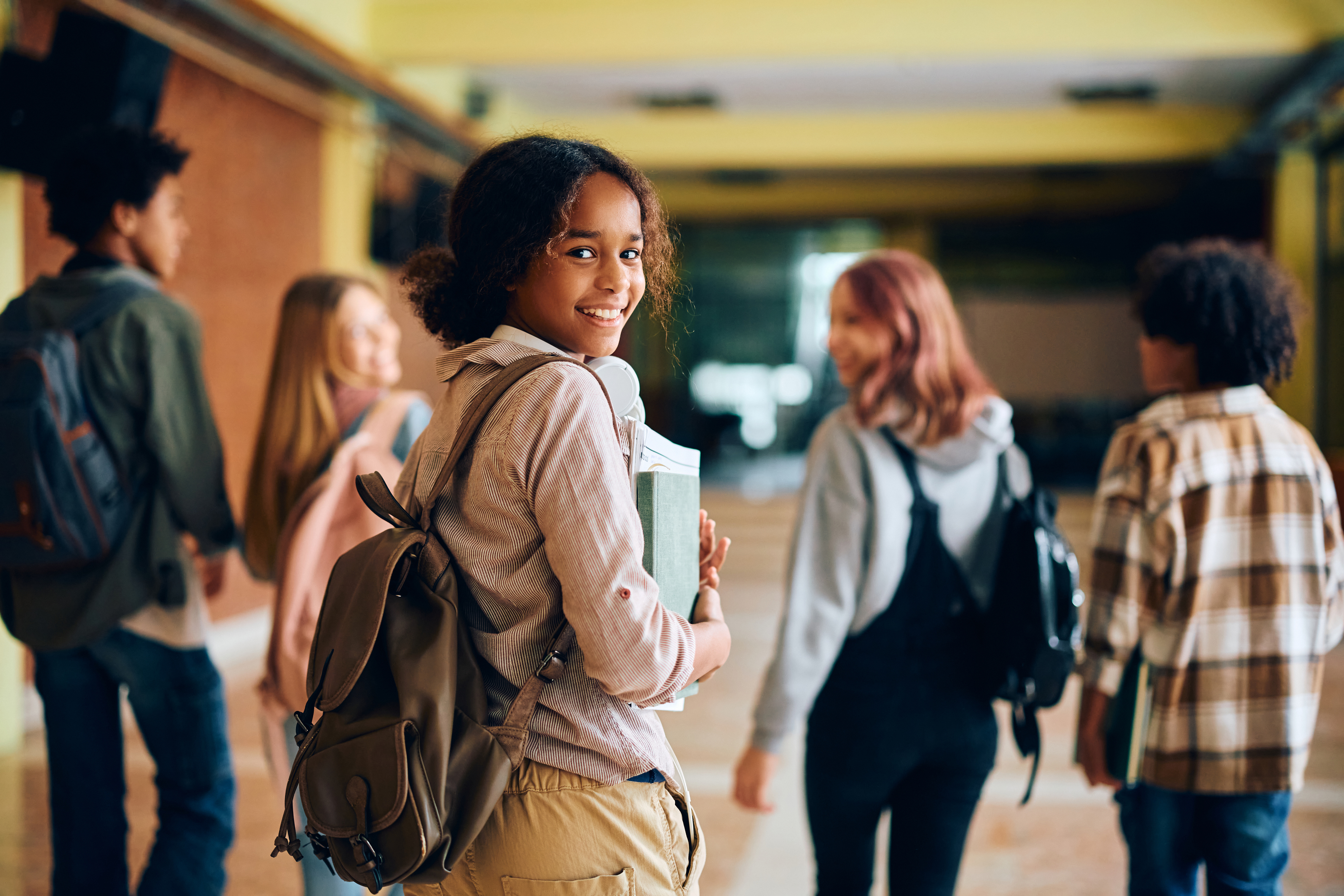 A group of five students walk down a school hallway. Focus is on a Black student with shoulder length curly hair; they are wearing a backpack and holding their school books smiling at the camera. The rest of the students are walking ahead and are out of focus. 