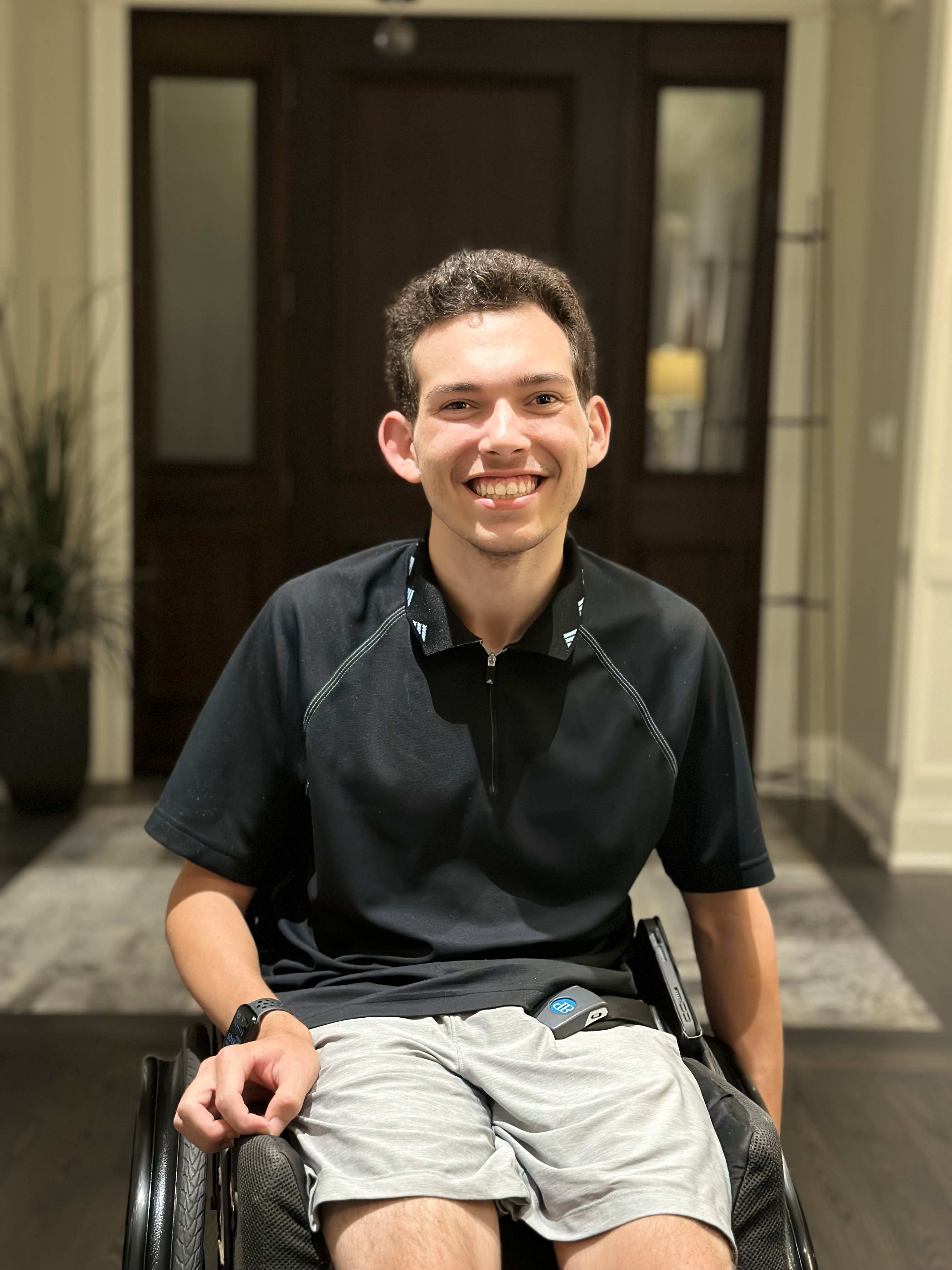 A portrait of Jake Wttewaal, a white young man with short dark hair. He is smiling, wearing a black polo shirt with grey athletic shorts. He is sitting in a wheelchair. 