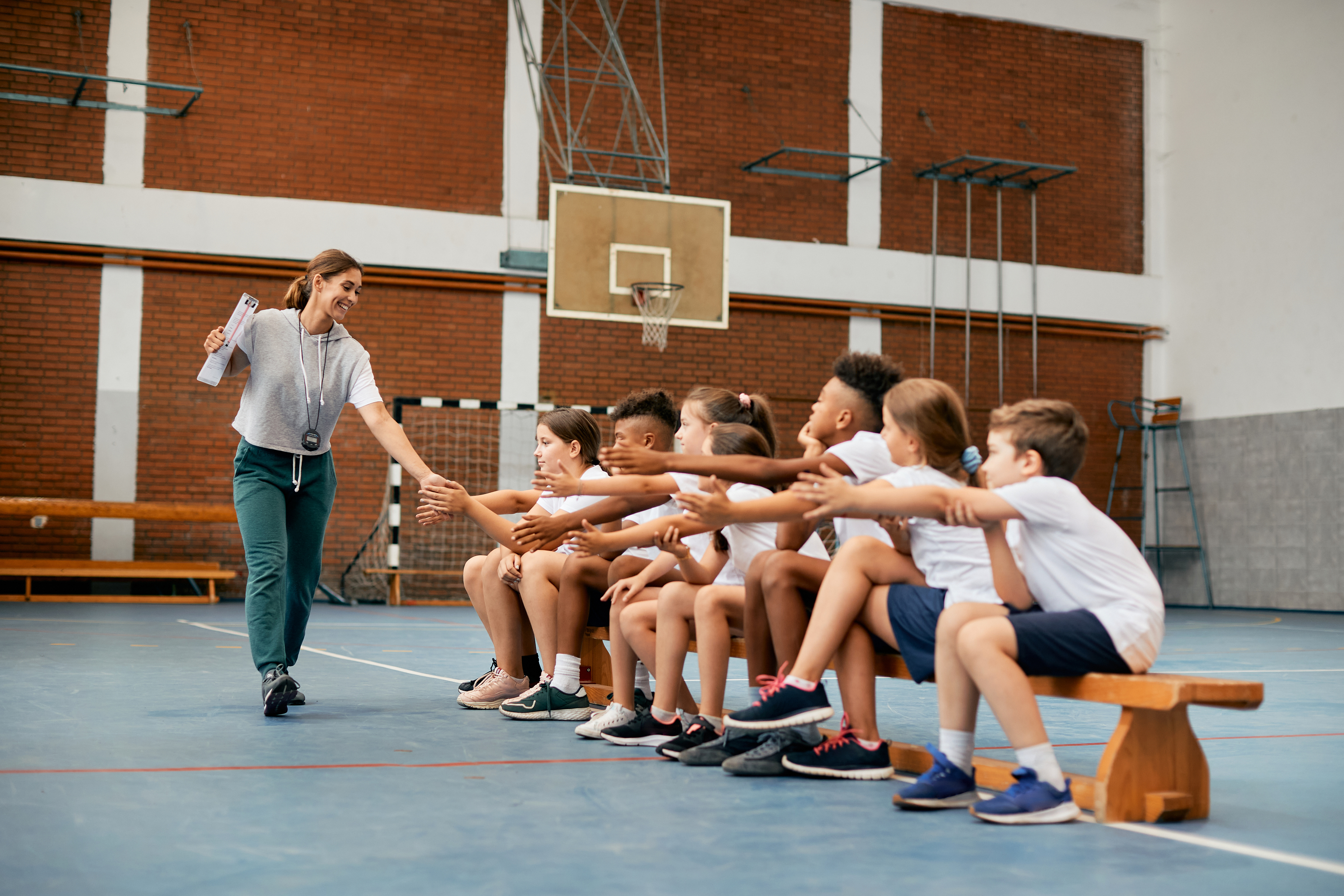A feminine-presenting gym teacher high fives a line of students extending their hands while sitting on a bench in a gymnasium. The teacher has long brown hair that is tied back in a ponytail, and is wearing athletic clothing and holding a clipboard. The students are diverse in terms of gender expression and skin tone. Everyone is smiling enthusiastically.