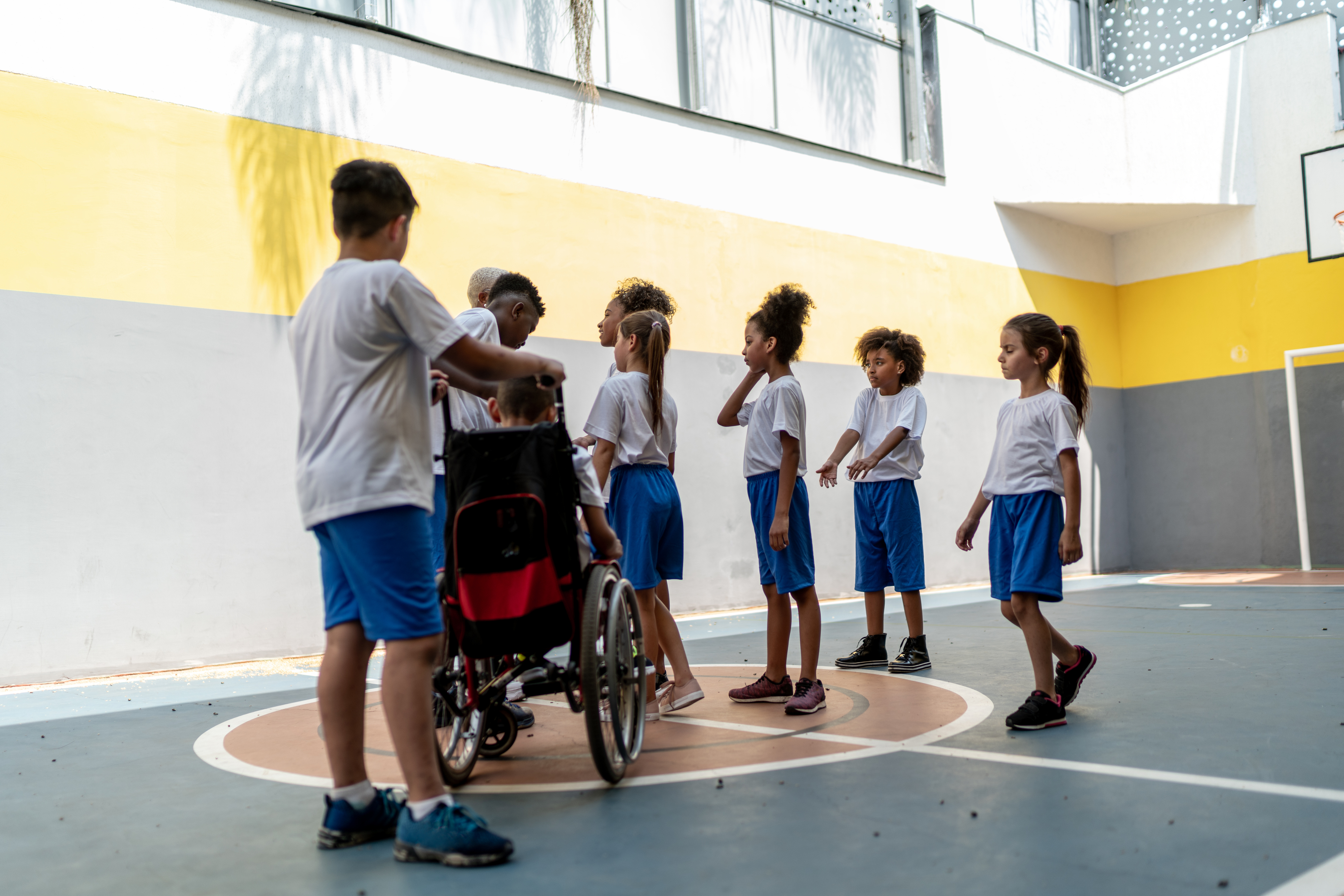 In a gymnasium, a diverse group of students wearing matching uniforms cluster around a teacher for instruction. The students display a range of gender presentations, skin tones, and hair textures. One of the students uses a wheelchair, and is being pushed by one of their peers to join the rest of the group. 