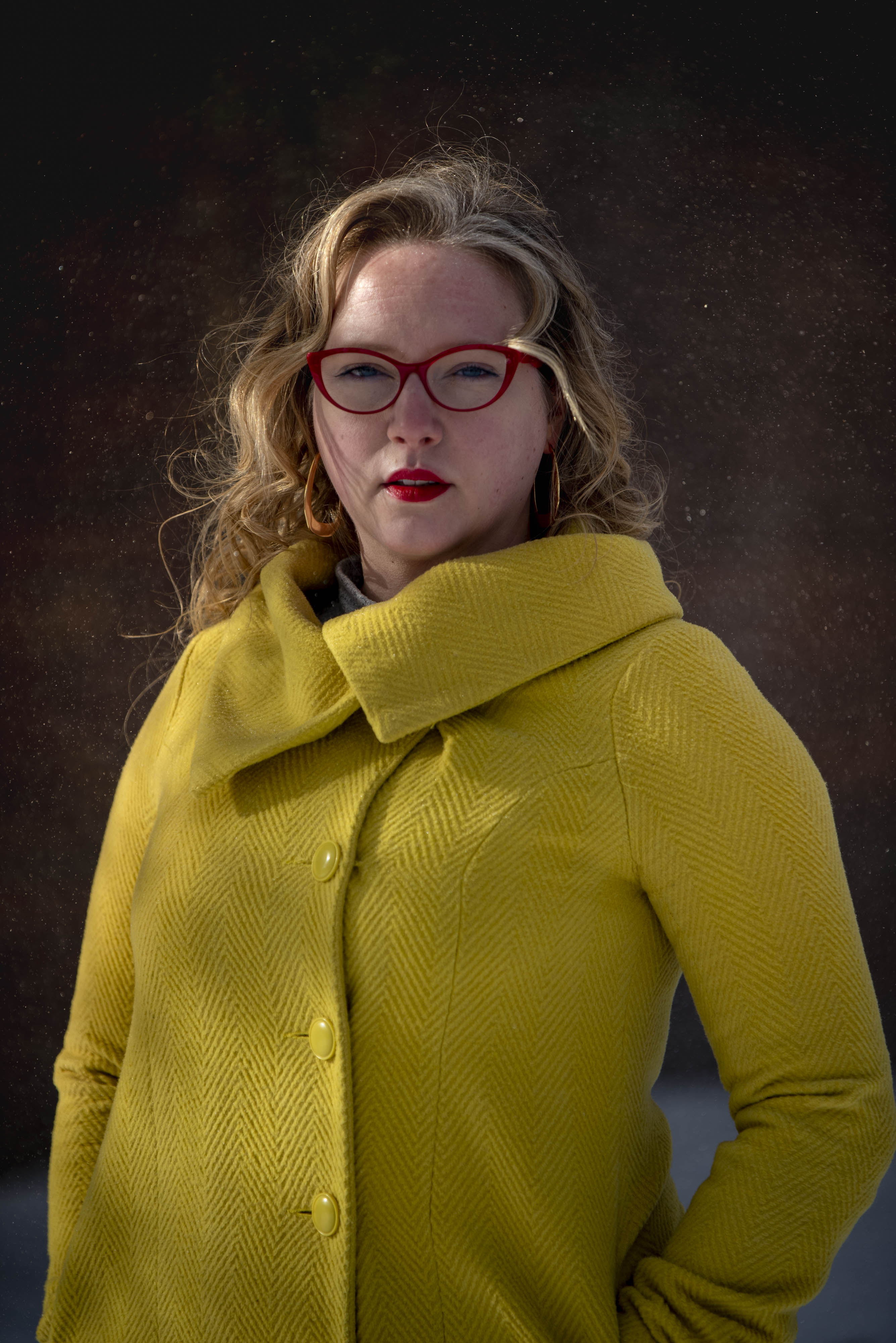 Une femme blanche aux longs cheveux blonds se tient à l'extérieur, regardant calmement la caméra. Elle porte des lunettes à monture rouge, du rouge à lèvres rouge et un manteau à chevrons jaune vif. 