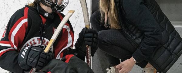 Jennifer Gallupe Roos, une femme blanche aux cheveux blonds, est agenouillée devant la porte ouverte d'un banc dans un stade de hockey, à côté d'un jeune athlète dans une luge de hockey. Jenn porte un chapeau noir et une veste noire, tandis que l'élève porte un casque et un équipement rouge et noir. 