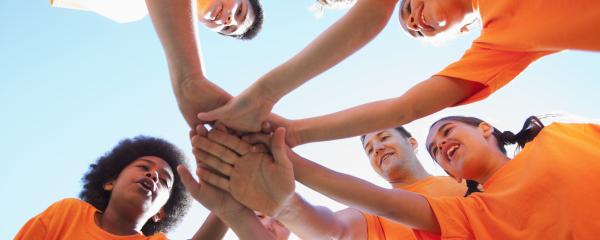 A low-angle image of six youth and their coach, wearing matching orange t-shirts, standing in a circle and stretching their hands towards the middle. The camera is positioned under the pile of hands. The youth display a variety of skin tones, hair colours and textures, and gender presentations.  