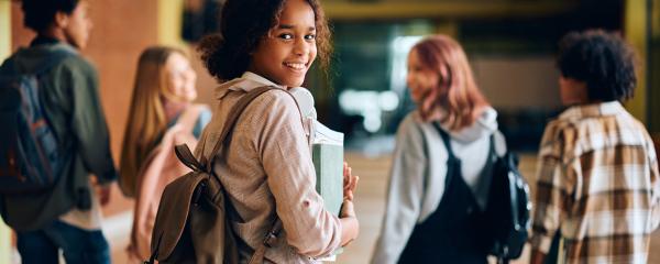 Cinq élèves marchent dans un couloir d’école. L’objectif est sur une élève noire aux cheveux bouclés mi-longs qui porte un sac à dos, tient ses manuels scolaires et sourit à l’appareil photo. Les autres élèves marchent devant et sont flous.