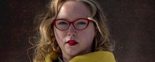 A white woman with long blonde hair stands outdoors, looking calmly at the camera. She is wearing red-rimmed glasses, red lipstick, and a bright yellow herringbone coat.  