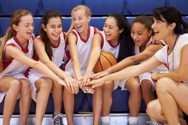 A youth basketball team sits on blue bleachers. Their hands are extended inwards to form a pile and they are smiling at one another. All of the players have long hair secured into a ponytail.
