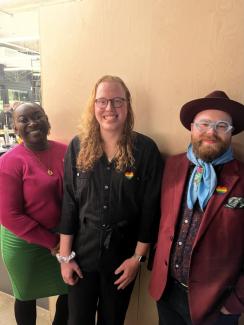 ""Three individuals stand smiling at the camera in front of a neutral background. The first from the left is a dark-skinned Black person wearing a bright fuschia top, green skirt, and yellow accessories. The person in the middle has long red curly hair and wears glasses and a black jumpsuit decorated with a heart-shaped sticker featuring the Pride flag. The person on the right of the image wears a maroon suit complete with matching hat and also decorated with a heart-shaped Pride flag sticker, a bright blue