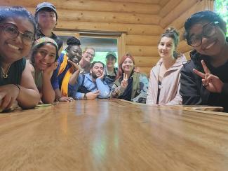 ""Ten people sit grouped around the far end of a wooden table. All are smiling at the camera, and some are making peace signs with their hands. The group is diverse in terms of racial background and gender expression. The photo was taken in a log cabin, and glimpses of green foliage are visible through the windows that aren’t obscured by people leaning in for the photo. 