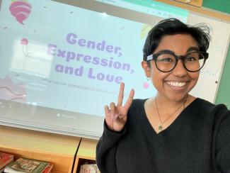 One individual smiling at the camera in front of a presentation screen reflecting a title slide reading Gender, Expression and Love. The person is wearing glasses and is making the peace sign with their hands. 