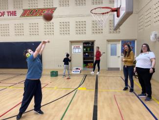 A student wearing athletic clothing and large noise-cancelling headphones is in the final stages of completing a basketball shot. The ball is in their air above them as they complete the follow-through.