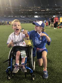 Two young children proudly display medals to the camera at the edge of a soccer field. One of the children uses a wheelchair, and the other kneels beside them. Both children appear excited about their achievement!