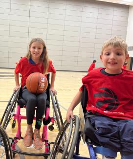 Two students using wheelchairs smile at the camera. They are in a gymnasium, and one of the students holds a basketball in their lap. 