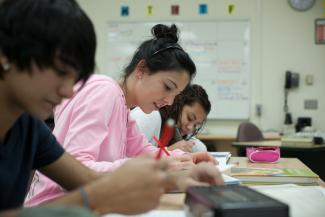 Close-up of three students working at their desks. The only student in focus wears their long dark hair in a bun and a pink sweater. 