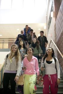 A group of laughing students walks down a wide flight of stairs towards the camera. All students are dressed casually, carrying backpacks and other school supplies. 