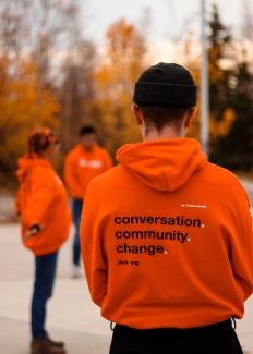 Three people stand on an outdoor basketball court with an autumn background. A person with short hair wearing a beanie stands with their back to the camera, displaying the text on the back of their orange Jack.org hoodie: “conversation. community. change.” Two out of focus individuals wear the same hoodie.