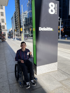 Jake Wttewaal, un jeune homme blanc aux cheveux bruns, sourit et montre un badge d'accès à côté d'un immeuble de bureaux. Il est assis dans un fauteuil roulant et porte une chemise bleue à rayures, un pantalon foncé et des chaussures noires.