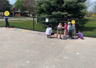 A group of students stand in front of a row of hula hoops that have been placed on the ground. One student is tossing an object towards the hula hoops while the rest observe and wait their turn. 