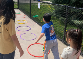 A group of students stand in front of a row of hula hoops that have been placed on the ground. One student is tossing an object towards the hula hoops while the rest observe and wait their turn. 