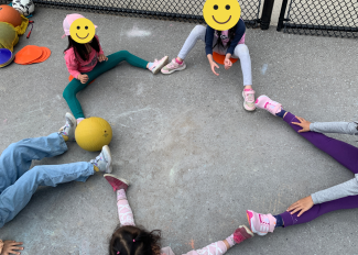Group of five students sitting on the ground. Their legs stretched out and connect to form the shape of a star.
